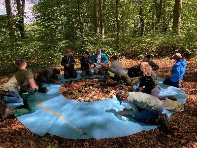 10 Jugendliche sitzen mit zwei Erwachsenen auf einer blauen Plane auf einem Waldboden und unterhalten sich