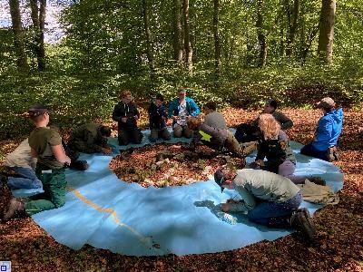 10 Jugendliche sitzen mit zwei Erwachsenen auf einer blauen Plane auf einem Waldboden und unterhalten sich